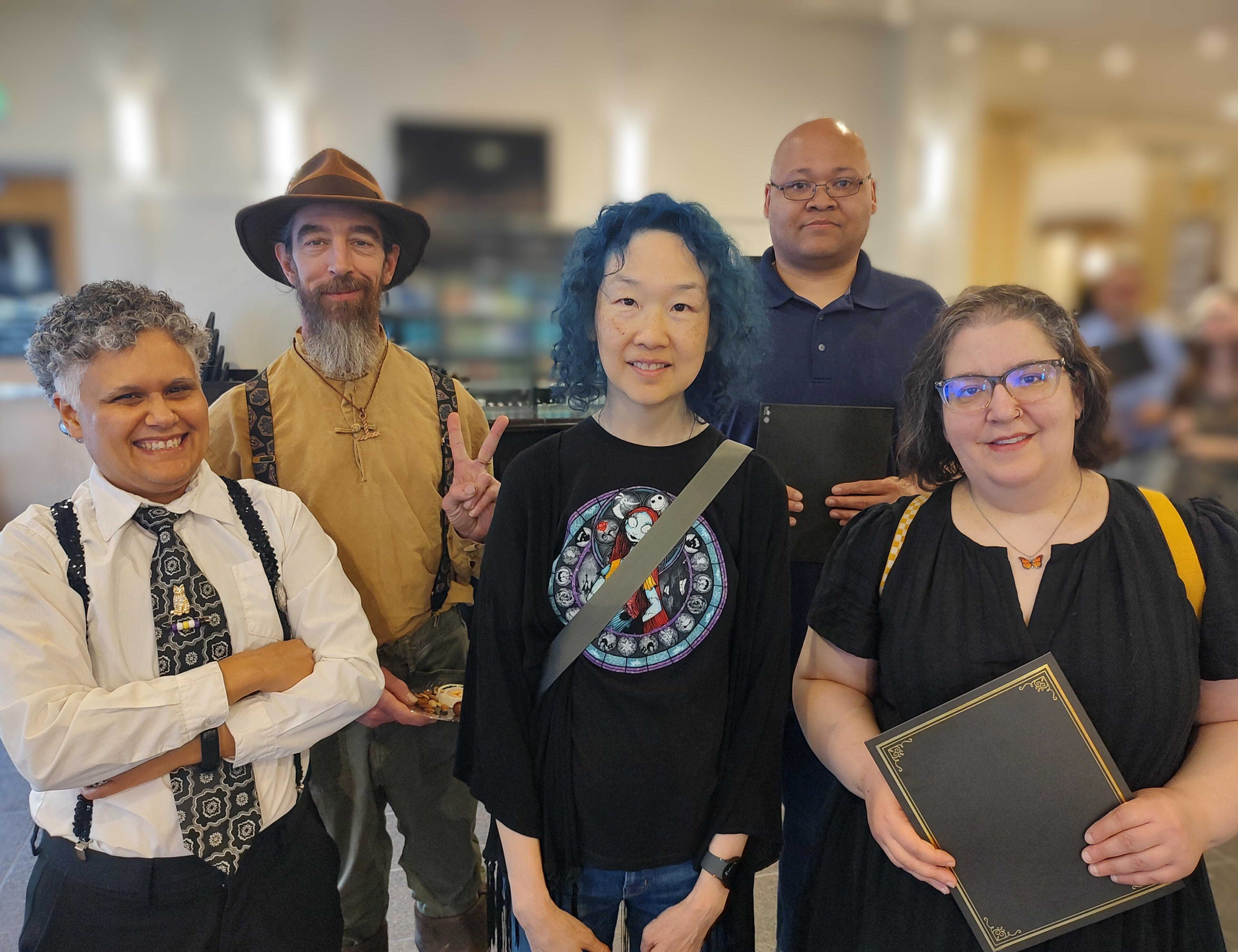 Velvet Rasputin LLC - collective members Vas is an Afro-Taíno-Celtiberian Puerto Rican person with short curly and gray hair. Loki is a bearded man of German and Central European descent, with long hair and a hat. Both are wearing long sleeve button-down shirt and suspenders. Chinyin is a Chinese